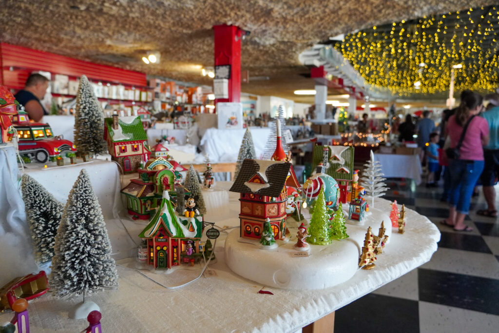 Festive miniature Christmas village display inside the Shell Factory & Nature Park in Fort Myers, Florida. The exhibit features detailed holiday-themed buildings, snow-covered trees, and twinkling lights, creating a winter wonderland atmosphere. Visitors browse the large indoor market filled with seasonal decorations, collectibles, and unique gifts.