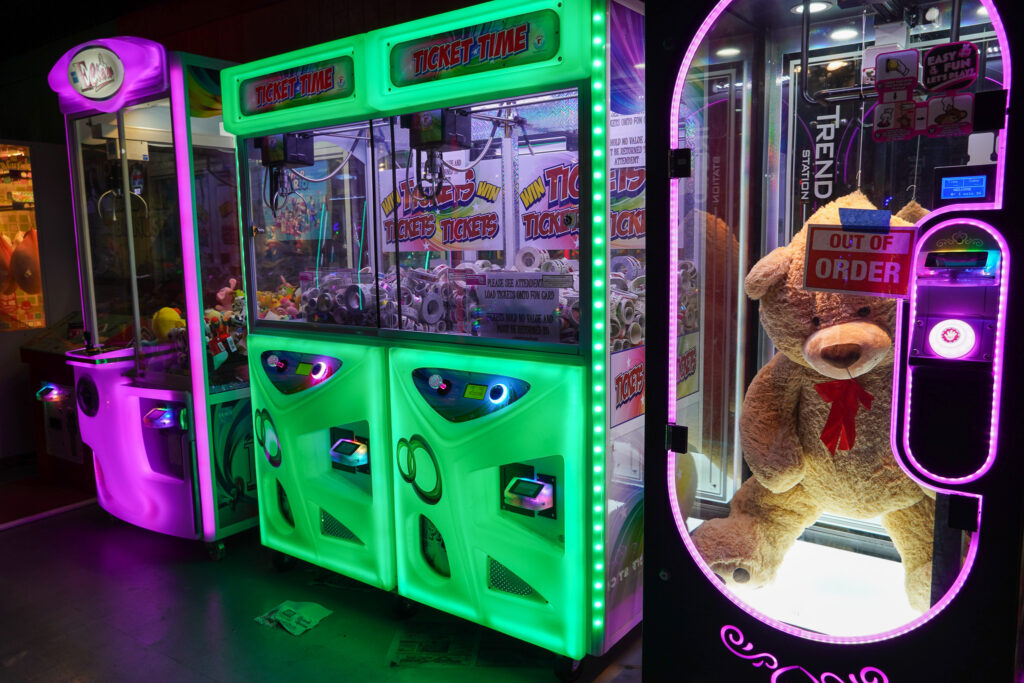 "A row of illuminated arcade claw machines at the Shell Factory & Nature Park in Fort Myers, Florida. The machines glow in vibrant shades of green, purple, and pink. One machine, labeled 'Ticket Time,' is filled with rolls of arcade tickets. Another machine features a giant plush teddy bear but is marked 'Out of Order' with a sign taped to its glass. The dimly lit arcade setting enhances the neon lighting, creating a nostalgic gaming atmosphere.