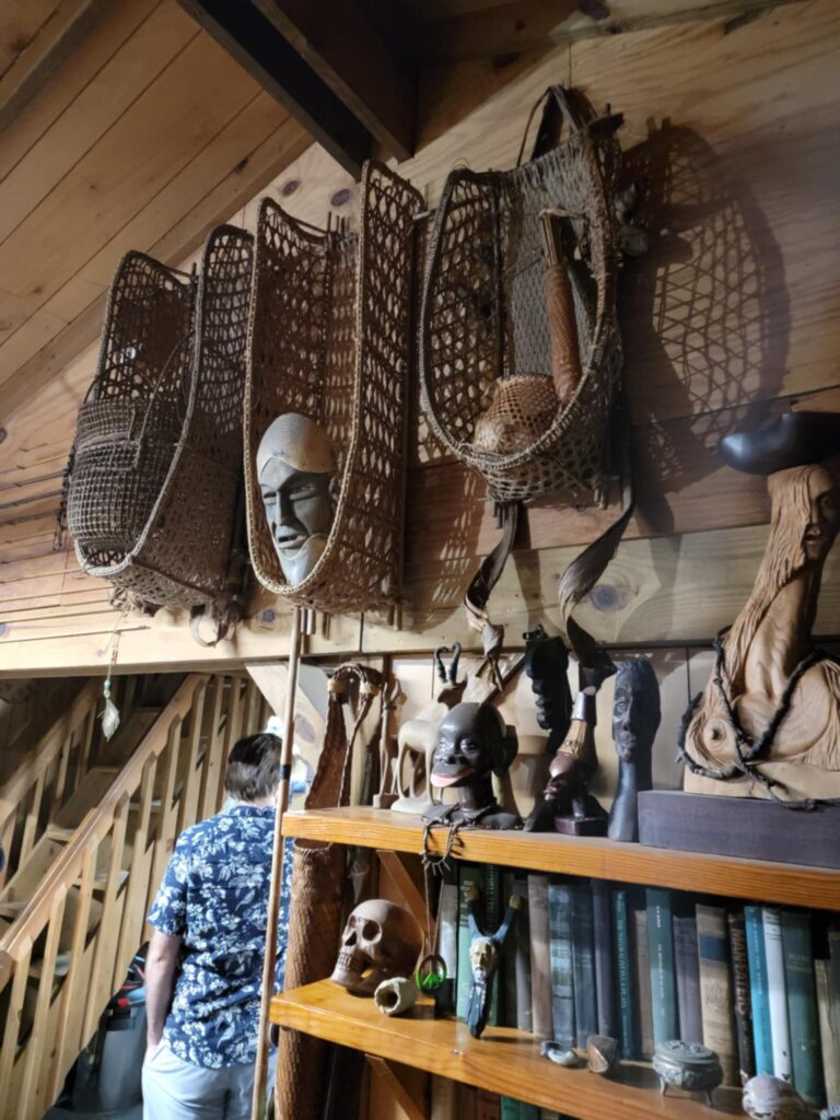 Inside the eclectic home of Sleeze Weazel in Gainesville, Florida, during his 75th birthday celebration. The rustic wooden interior features tribal masks, woven fish traps, carved wooden figurines, and antique books. A man in a Hawaiian shirt explores the space, adding to the mysterious and cultural ambiance of the collection. Unique artifacts and shadowed lighting create a museum-like atmosphere.