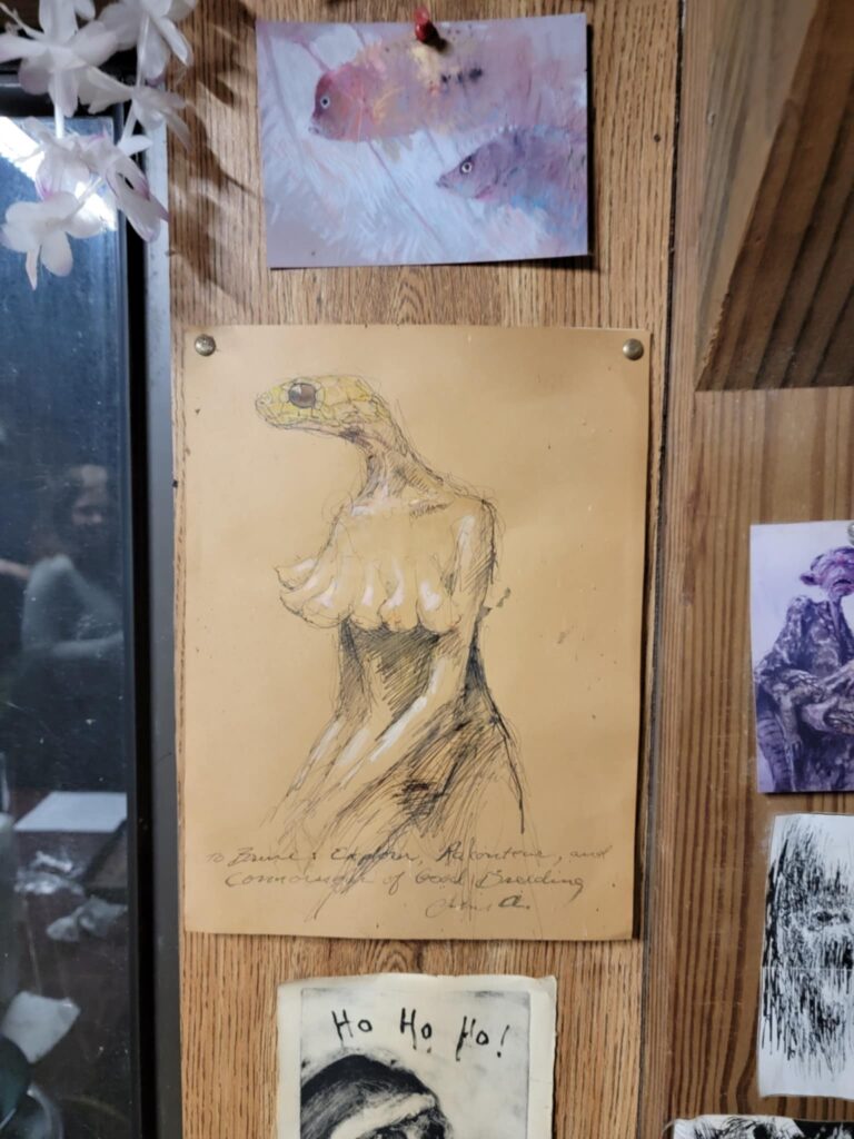 Unusual artwork displayed at Sleeze Weazel's house in Gainesville, Florida, during his 75th birthday celebration. The focal point is a hand-drawn sketch of a humanoid reptilian figure with exaggerated human features, pinned to a wooden wall. Surrounding the sketch are other eclectic artworks, including a pastel painting of fish and a dark, eerie holiday-themed image with 'Ho Ho Ho!' written on it. The collection reflects a mix of surrealism, folklore, and outsider art.