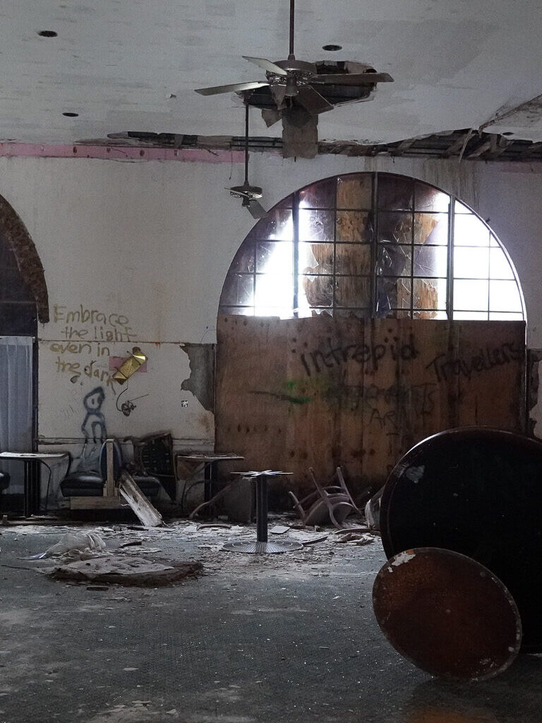 Interior of the abandoned Port of the Islands Hotel in Naples, FL, prior to its demolition. The room features large arched windows, partially boarded up, with ceiling fans hanging from a damaged ceiling. Graffiti-covered walls and debris-strewn floors, including broken furniture and overturned tables, reflect the state of decay. This haunting scene encapsulates the eerie atmosphere of abandonment and neglect.
